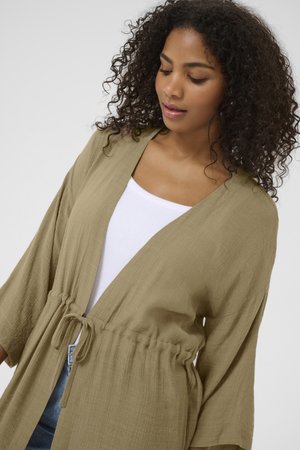 Olijfgroen, lichtgewicht kimono-stijl cardigan met een trekkoord in de taille, lange mouwen en een textuurstof, gelaagd over een witte top.