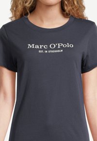 Marinblå bomulls T-shirt med kort ärm, med vit text "Marc O’Polo" och "EST. I STOCKHOLM" i ett modernt typsnitt centrerat på bröstet.