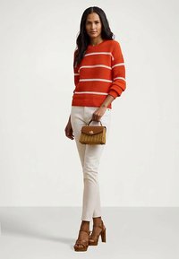 Mulher vestindo um suéter listrado em laranja e branco, calças brancas, sandálias de salto alto marrons, segurando uma pequena mala marrom com alça superior.