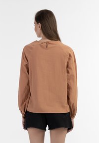 Langærmet top i rustorange stof med en tekstureret finish. Har en nøglehulslukning bagpå og en afslappet pasform, parret med sorte shorts.