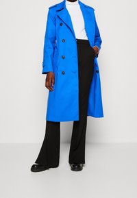 Blå trenchcoat med dubbelknäppt design, stora svarta knappar och axelapplikationer, i kombination med svarta vida byxor och svarta skor.