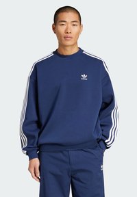 Mornarsko plava dukserica s bijelim trostrukim trakama na rukavima, okrugli izrez i izvezeni adidas logo na prsima. Mekana tekstura tkanine.