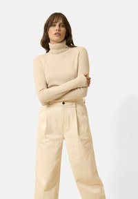 Maglione dolcevita beige realizzato in tessuto morbido, abbinato a pantaloni a gamba larga color crema a vita alta con dettagli di bottoni.