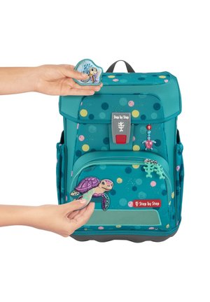 Hands attaching colorful turtle patches to a teal children's backpack with multi-colored dots and "Step by Step" label.