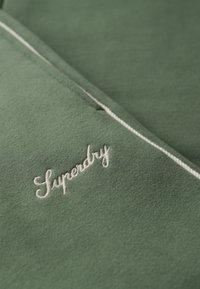 Grüner Stoff mit einer glatten Textur, der das weiße gestickte "Superdry"-Logo zeigt. Weiße Nähte sorgen für einen Kontrast im Design.