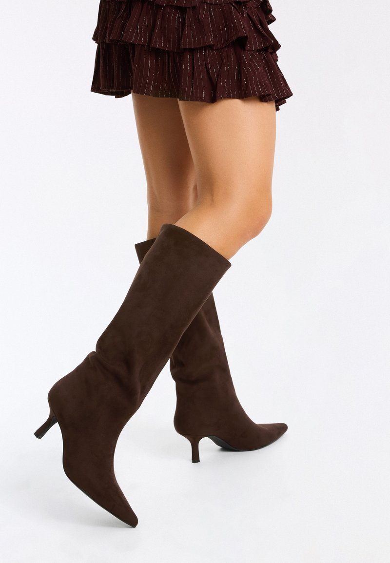 Personne portant des bottes à talons marron foncé montant jusqu’aux genoux et une jupe courte marron à volants avec des rayures métalliques, debout devant un fond uni.