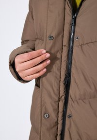 Doudoune marron avec une fermeture éclair sur le devant, des boutons-pression et une texture matelassée. Une main est visible près de l'ouverture, mettant en valeur le design.