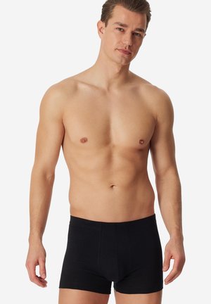 Shirtless man with short brown hair wearing fitted black boxer briefs, standing against a plain light background.
