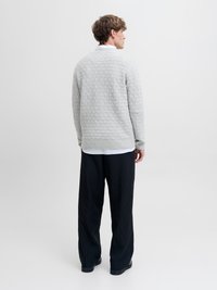 Άνδρας με την πλάτη στραμμένη φοράει ανοιχτό γκρι υφασμάτινο πουλόβερ, λευκό πουκάμισο, μαύρο παντελόνι με ρίγες και φαρδύ πόδι, και μαύρα παπούτσια σε απλό φόντο.