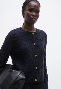 Καρντιγκάν ναυτικού μπλε με πλεκτό σχέδιο, στρογγυλή λαιμόκοψη και έξι χρυσά διακοσμητικά κουμπιά. Το μοντέλο κρατάει μια μαύρη τσάντα, παρουσιάζοντας υφή υφάσματος.