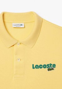 Camisa polo de algodón amarilla con cuello de punto, que presenta un cierre de dos botones y un logo de Lacoste en verde con un cocodrilo bordado.