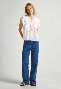 Blusa bianca di cotone con maniche a volant, scollatura con lacci e dettagli con nappe. Abbinata a jeans in denim blu a vita alta.