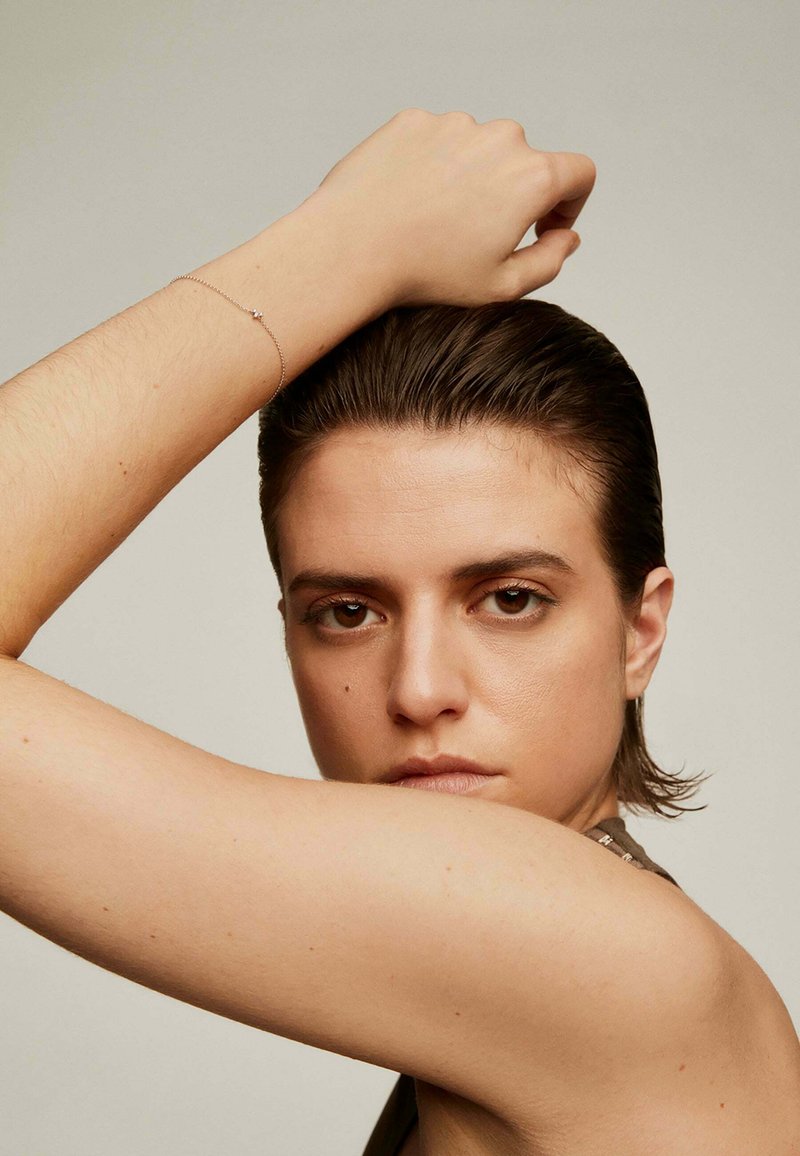 Pulsera de plata en la muñeca, con un diseño minimalista y un único elemento decorativo. Cabello peinado hacia atrás, fondo neutro.