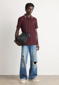 Bordeaux polo shirt met gestreepte kraag, blauwe distressed wijde jeans en crème schoenen. Het model houdt een zwarte leren clutch vast.