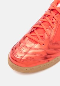 Sneaker sintético laranja com acabamento brilhante, sobreposições texturizadas e sola de borracha natural. Apresenta atacadores e perfurações para ventilação.