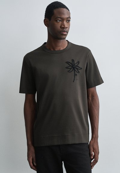 T-shirt vert olive à manches courtes en coton, avec un motif de fleur brodé en noir sur la poitrine gauche. Coupe simple et décontractée.