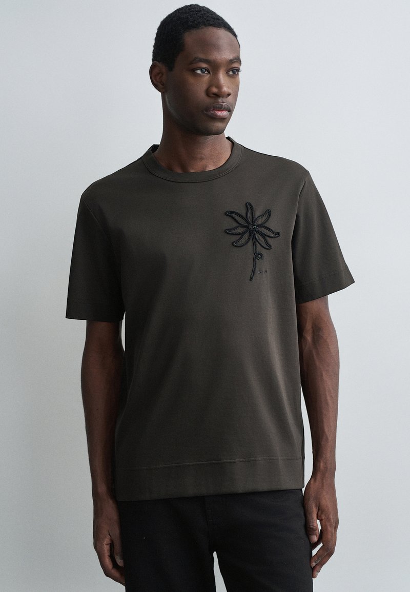 Tricou scurt cu mânecă, de culoare verde măslin, fabricat din bumbac, având un design cu floare brodată, de culoare neagră, pe partea stângă a pieptului. Croială simplă, lejeră.