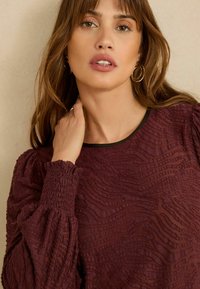 Burgundi texturált felső ráncolt puffadt ujjakkal, fekete szegélyű nyakkivágással és a teljes anyagra hullámos mintákkal.