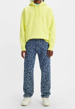 Personne portant un sweat à capuche jaune fluo, un jean bleu à motifs et des baskets blanches, debout devant un fond clair uni.