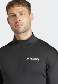 Pullover termico nero con collo a mezza zip, caratterizzato da dettagli grigi e un logo bianco "TERREX" sul lato sinistro del petto. Tessuto morbido e elasticizzato.