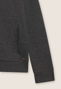 Pull long de couleur gris foncé à manches longues, avec une texture côtelée, une fente sur le côté, des détails aux poignets et une petite étiquette de marque marron près de l'ourlet.