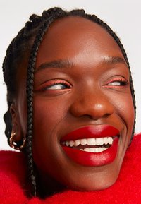 Close-up of a face with dark skin, braided hair, bright red lipstick, and soft red clothing. Features include golden earrings and subtle eye makeup.
