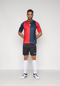 Koszulka piłkarska z krótkim rękawem w czerwono-granatowym, podzielonym wzorze z logo, w zestawie z czarnymi szortami i białymi, sięgającymi kolan skarpetami.