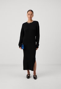 Črna rebrasta pletena obleka z dolgimi rokavi, stranskim razporkom in okroglim izrezom. Uporabljena je s črnimi čevlji in majhno modro torbico.