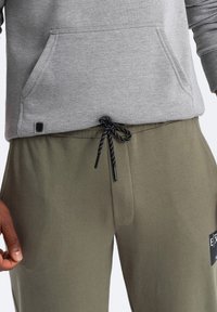 Grauer Hoodie mit einer Vorderasstasche, kombiniert mit olivgrünen Hosen mit einem schwarzen Kordelzug und einem Etikett am Bund. Glatte Textur.