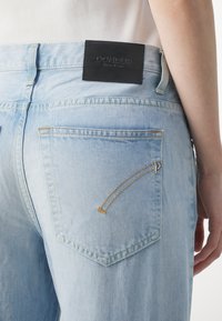 Vaaleansiniset farkut, joissa on kulutettu viimeistely, musta nahkainen merkkilaatta, oranssit saumat ja vetoketjullinen takatasku.