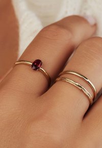 Des bagues en or avec une finition lisse et une avec une pierre précieuse rouge ovale. Trois bagues empilées sur une main ayant une apparence de peau texturée.