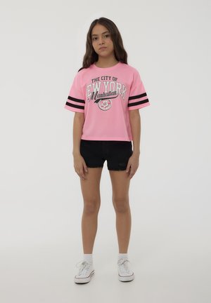 T-shirt cropped rosa con strisce nere sulle maniche e grafica con il testo "LA CITTÀ DI NEW YORK Manhattan." Abbinate a pantaloni corti di denim neri e sneakers bianche.