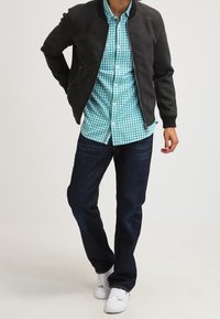 Chaqueta bomber negra, camisa a cuadros turquesa, vaqueros oscuros y zapatillas blancas. La chaqueta tiene bolsillos con cremallera. Conjunto casual.