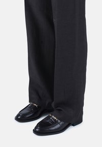 Black leather loafers with textured detailing, featuring a gold accessory at the front, paired with wide, dark trousers.