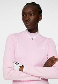 Ljusrosa sportjacka med en texturerad design, framside dragkedja och vita detaljer. Har en vit logotyp på bröstet och kontrasterande ärmslut.