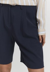 Pantalones cortos azul marino a medida con una textura suave, con pliegues frontales y bolsillos laterales. Combinados con una camisa a rayas beige y azul.