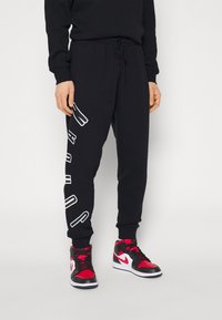 Pantalones deportivos de algodón negros con cintura elástica y cordón, con gráficos de texto en blanco en la pierna izquierda. Combinados con zapatillas rojas y negras.