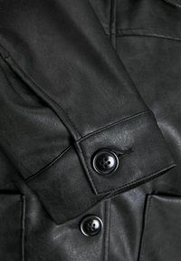 Manche de veste en cuir noir avec une fermeture à bouton. Présente une texture lisse et un détail de couture prononcé près de l'épaule.