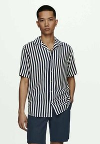 Κοντομάνικο πουκάμισο με κουμπιά, σε ναυτικό και λευκό κάθετο ρίγα, κλασικό γιακά και χαλαρή γραμμή. Φορεμένο με ναυτικά σορτς.