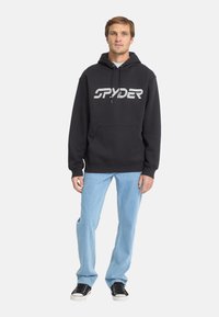 Felpa nera con logo "SPYDER" in strisce bianche, tasca a marsupio, cordini regolabili, abbinata a jeans azzurri chiari e sneakers nere.