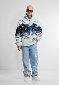 Fleecegenser i lys blå med palmer og tigre trykt i svart og blått; kombinert med slitte, lyseblå jeans.