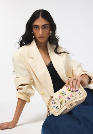 Vrouw met lang donker haar, gekleed in een crèmekleurig colbert en zwarte top, houdt een beige tas met bloemrijke borduursels en gouden ketting vast tegen een witte achtergrond.