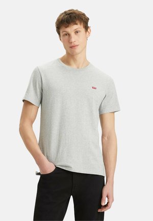 Ung mand iført en lysegrå Levi's t-shirt og sorte bukser, stående med den ene hånd i lommen op ad en ensfarvet baggrund.
