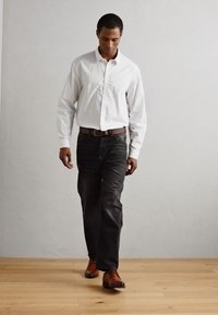 Camicia a maniche lunghe a righe bianche, jeans scuri, cintura in pelle marrone e scarpe marroni. La persona sta camminando su un pavimento di legno contro un muro chiaro.