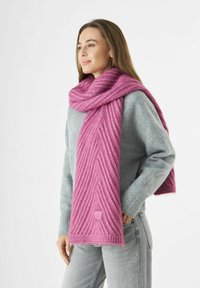 Vrouw in een dikke roze gebreide sjaal over een grijze trui en lichtgrijze jeans, staande met één hand in haar zak, kijkend naar de zijkant.