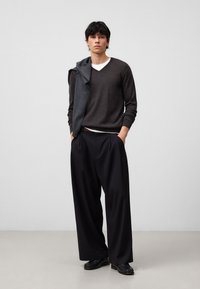 Dunkelbrauner Strickpullover über einem weißen T-Shirt, kombiniert mit hoch taillierten schwarzen Hosen. Schwarze Schuhe ergänzen den lässigen Schnitt und die überschnittenen Schultern.