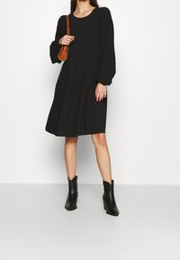 Černé šaty s dlouhým rukávem a vrstevnatým designem, s elastickými manžetami. Nosené s černými botami po kotníky a hnědou crossbody kabelkou.