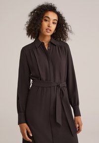 Robe chemise noire à ceinture, avec des manches longues, un front à boutons et des épaules froncées. Texture douce avec une finition lisse. Pas de motifs distincts ni de matériel.