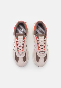 Zapatillas Adidas con una parte superior de malla en un ligero tono beige y marrón, detalles en rojo y rayas blancas, con una suela de goma texturizada y cierre de cordones.