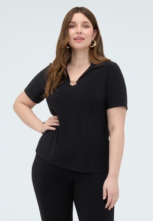 Kvinde med langt brunt hår iført guldringe i ørerne, sort kortærmet top med krave og sorte bukser, poserer med hånden på hoften.